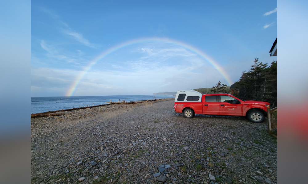 Véhicule rouge de RNCan garé sur une plage rocheuse avec un arc-en-ciel en arrière-plan